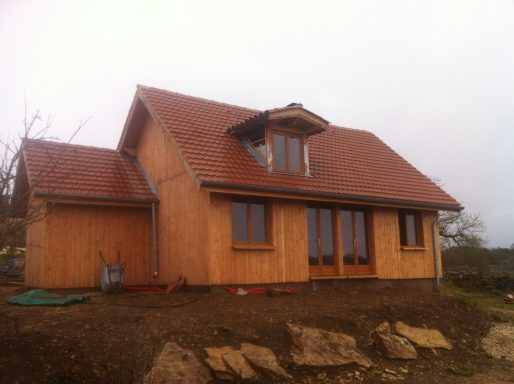 Maison en bois à deux étages avec toit en tuiles, entourée de terrain nu.