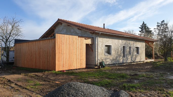 Maison en Paille avec une extension en bois, entourée de terrain et ciel dégagé.