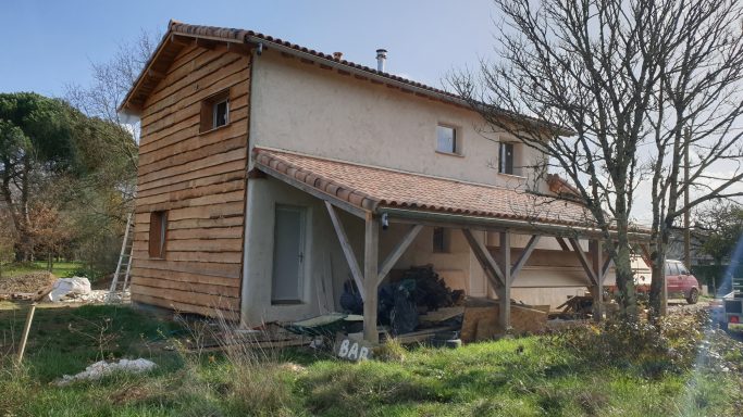 Maison à deux niveaux en bois avec un porche, entourée de verdure.
