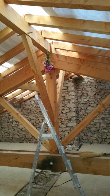 Poutres en bois dans un grenier, échelle en métal et mur en pierres apparentes.