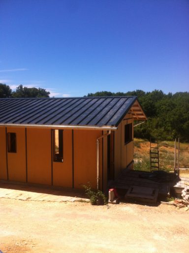 Maison moderne en construction avec un toit noir sous un ciel bleu, entourée de verdure.