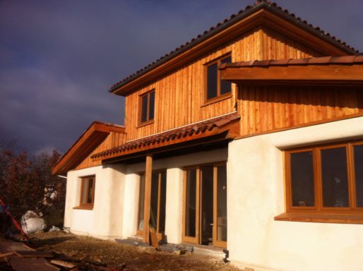 Maison à deux étages avec façade en bois et fenêtres en bois, sous un ciel nuageux.