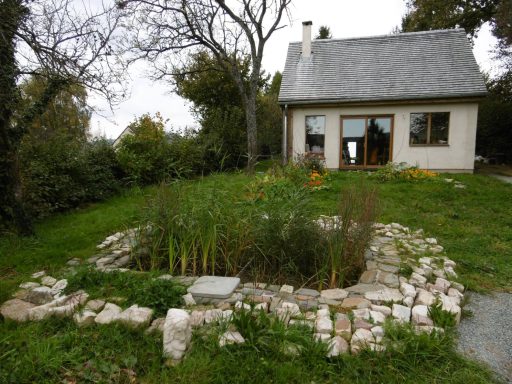 Maison avec un toit en pente, entourée d'un jardin et d'un étang de plantes.